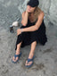Woman sitting on a rocky surface wearing a black dress and blue sandals.